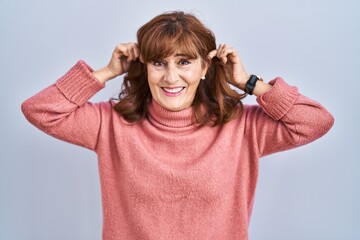 Middle age hispanic woman standing over isolated background smiling pulling ears with fingers,...