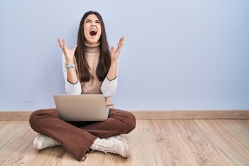 Young brunette woman working using computer laptop sitting on the floor crazy and mad shouting and yelling with aggressive expression and arms raised. frustration concept.