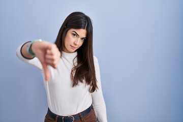 Young brunette woman standing over blue background looking unhappy and angry showing rejection and negative with thumbs down gesture. bad expression.