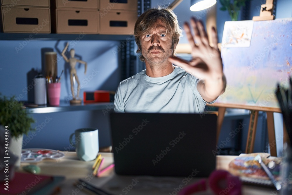 Poster Middle age man sitting at art studio with laptop at night doing stop sing with palm of the hand. warning expression with negative and serious gesture on the face.