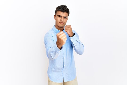 Young hispanic man wearing business shirt standing over isolated background ready to fight with fist defense gesture, angry and upset face, afraid of problem