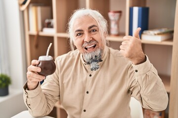 Middle age man with grey hair drinking mate infusion pointing thumb up to the side smiling happy with open mouth