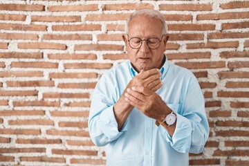 Senior man with grey hair standing over bricks wall suffering pain on hands and fingers, arthritis...