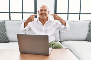 Senior man using laptop at home sitting on the sofa smiling cheerful showing and pointing with fingers teeth and mouth. dental health concept.