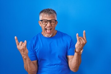 Naklejka premium Hispanic man with grey hair standing over blue background shouting with crazy expression doing rock symbol with hands up. music star. heavy music concept.