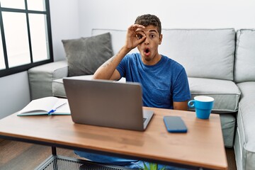 Young handsome hispanic man using laptop sitting on the floor doing ok gesture shocked with surprised face, eye looking through fingers. unbelieving expression.