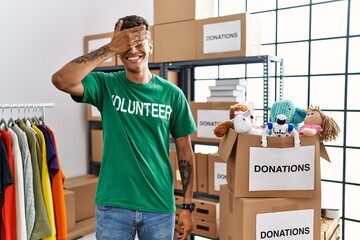 Young handsome hispanic man wearing volunteer t shirt at donations stand smiling and laughing with hand on face covering eyes for surprise. blind concept.