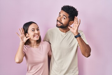 Young hispanic couple together over pink background smiling with hand over ear listening an hearing to rumor or gossip. deafness concept.