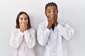 Young hispanic doctors standing over white background laughing and embarrassed giggle covering...