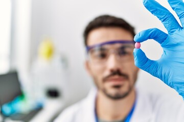 Young hispanic man wearing scientist uniform holding pill at laboratory
