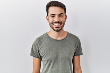 Young hispanic man with beard wearing casual t shirt over white background winking looking at the camera with sexy expression, cheerful and happy face.