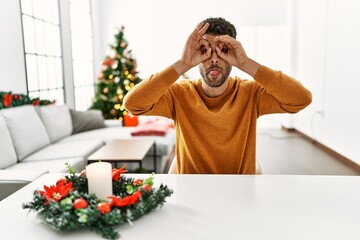 Arab young man sitting on the table by christmas tree doing ok gesture like binoculars sticking tongue out, eyes looking through fingers. crazy expression.
