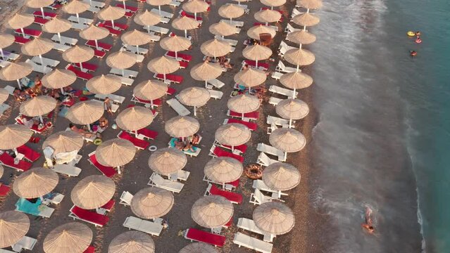 Beach beds and rows of thatch umbrellas - tiki straw parasols by the sea. Waves crashing on the sand (lapping at the shore). Aerial drone view on the coast at a summer vacation resort.