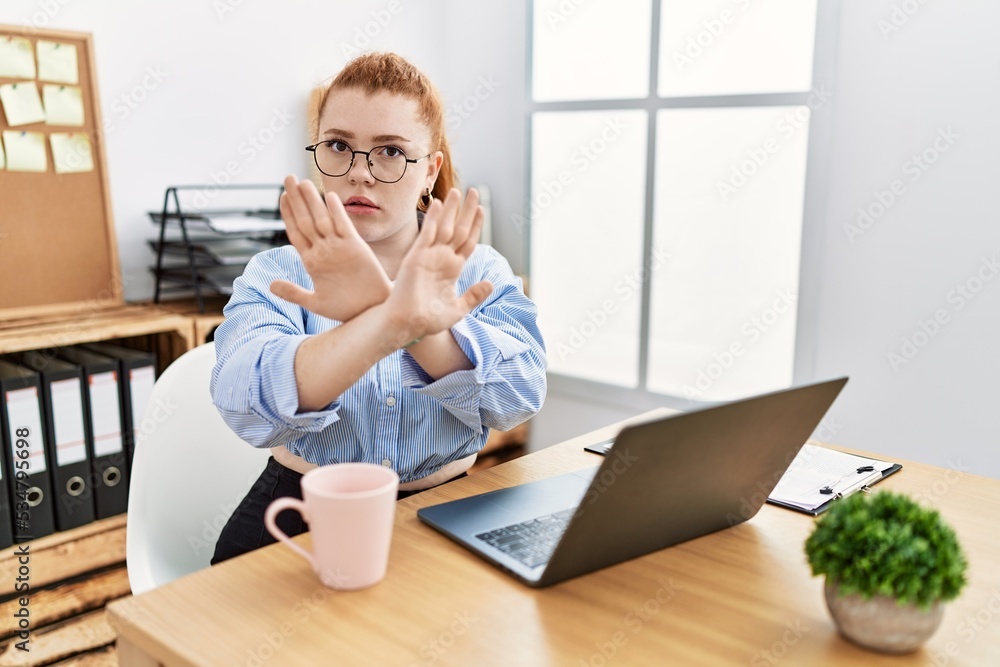 Wall mural Young redhead woman working at the office using computer laptop rejection expression crossing arms and palms doing negative sign, angry face