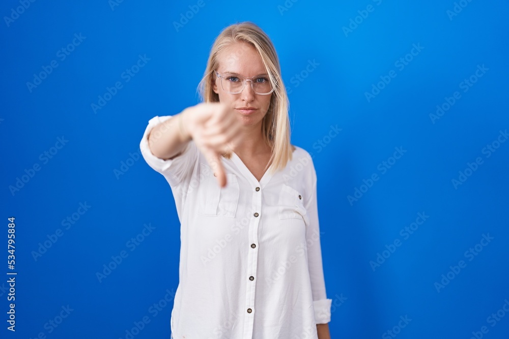 Sticker Young caucasian woman standing over blue background looking unhappy and angry showing rejection and negative with thumbs down gesture. bad expression.