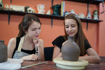 Close-up of concentrated beautiful craftswoman in apron sitting at pottery wheel and using craft tool while shaping wet clay vessel