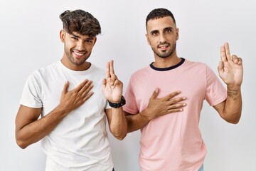 Young gay couple standing over isolated background smiling swearing with hand on chest and fingers...