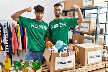 Young gay couple wearing volunteer t shirt at donations stand strong person showing arm muscle,...