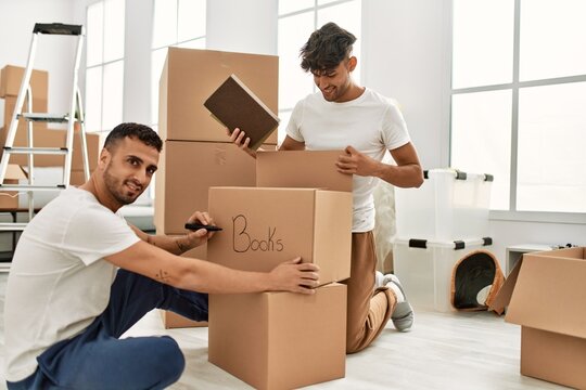 Two hispanic men couple smiling confident packing book cardboard box at new home