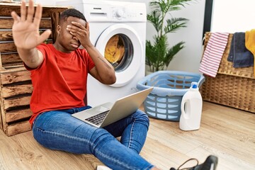 Young african man doing laundry and using computer covering eyes with hands and doing stop gesture...