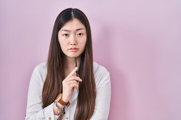 Chinese young woman holding pencil over pink background relaxed with serious expression on face. simple and natural looking at the camera.