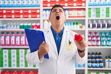 Young hispanic man working at pharmacy drugstore holding heart angry and mad screaming frustrated and furious, shouting with anger looking up.