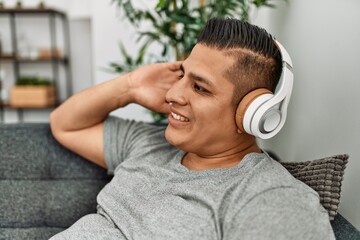 Young hispanic man smiling happy listening music sitting on the sofa at home.