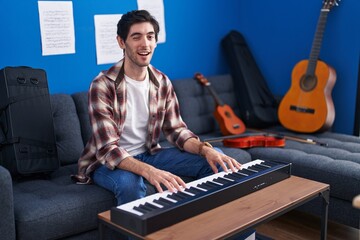 Young hispanic man playing piano at music studio winking looking at the camera with sexy expression, cheerful and happy face.