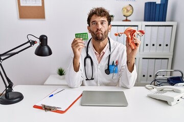 Young hispanic doctor man holding female genital organ and birth control pills making fish face with mouth and squinting eyes, crazy and comical. - Powered by Adobe