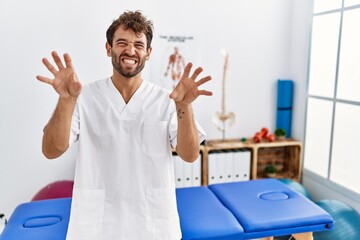 Young handsome physiotherapist man working at pain recovery clinic smiling funny doing claw gesture...