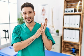 Young physiotherapist man working at pain recovery clinic clapping and applauding happy and joyful, smiling proud hands together