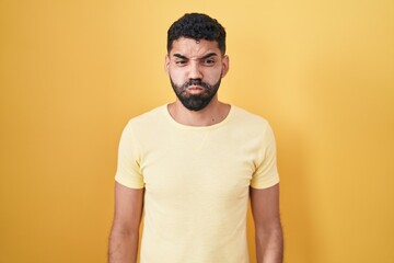 Hispanic man with beard standing over yellow background puffing cheeks with funny face. mouth inflated with air, crazy expression.