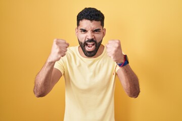 Hispanic man with beard standing over yellow background angry and mad raising fists frustrated and...
