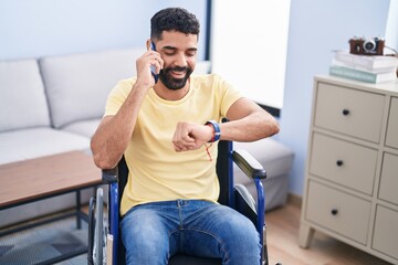 Young arab man talking on smartphone looking watch sitting on wheelchair at home