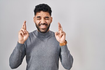 Hispanic man with beard standing over white background gesturing finger crossed smiling with hope...