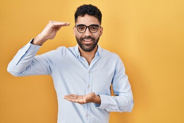 Hispanic man with beard standing over yellow background gesturing with hands showing big and large size sign, measure symbol. smiling looking at the camera. measuring concept.