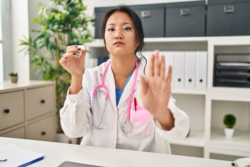 Young asian woman wearing doctor uniform and stethoscope with open hand doing stop sign with serious and confident expression, defense gesture