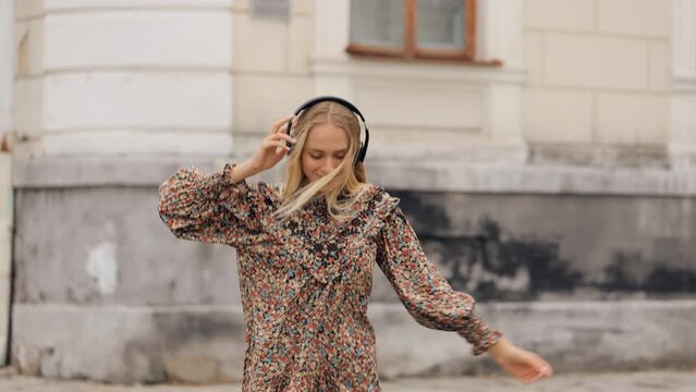 Slow motion of happy young long hair woman in headphones walking outdoors in city street having fun alone. Joyful attractive blonde carefree woman listening to music