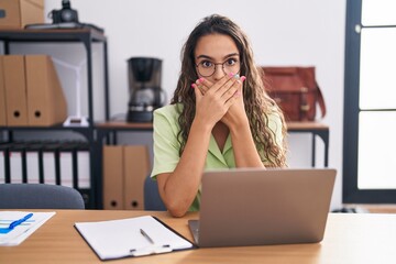 Young hispanic woman working at the office wearing glasses shocked covering mouth with hands for mistake. secret concept.