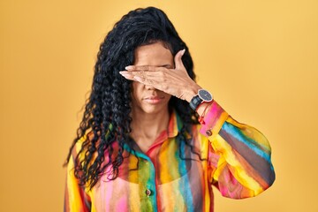 Middle age hispanic woman standing over yellow background covering eyes with hand, looking serious...