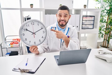 Hispanic doctor man holding clock at the clinic smiling happy pointing with hand and finger