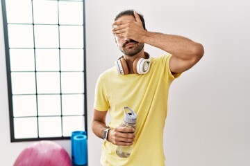 Young hispanic man wearing sportswear and drinking water at the gym covering eyes with hand,...