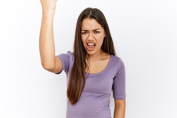 Young brunette woman standing by isolated background angry and mad raising fist frustrated and...