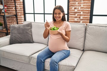Young latin woman pregnant eating salad sitting on sofa at home