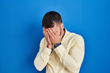 Handsome hispanic man standing over blue background with sad expression covering face with hands while crying. depression concept.