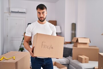 Handsome hispanic man moving to a new home holding fragile box skeptic and nervous, frowning upset...