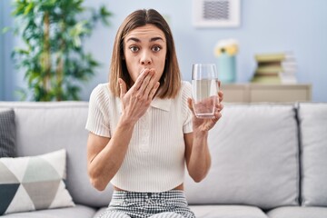 Hispanic woman drinking glass of water covering mouth with hand, shocked and afraid for mistake....