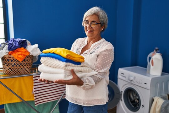 Middle age woman smiling confident holding folded clothes at laundry room