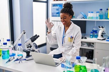Young african american with braids working at scientist laboratory with laptop doing ok sign with...