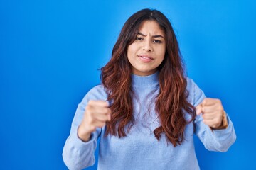 Fototapeta premium Hispanic young woman standing over blue background angry and mad raising fists frustrated and furious while shouting with anger. rage and aggressive concept.